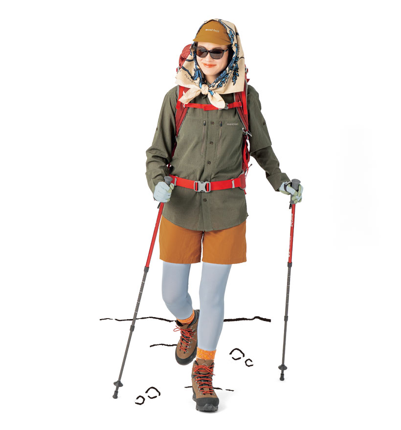 カーキの長袖シャツ、ブラウンのショートパンツ、白いタイツを着用した女性モデル。サングラスをかけ、茶褐色のキャップの上にベージュのスカーフをかぶり首元で巻いている。中型の赤いバックパックを背負い、赤いトレッキングポールを持っている