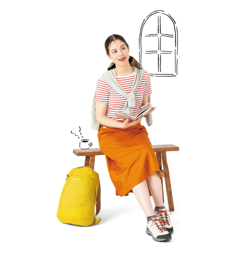 赤と白のボーダーTシャツと茶色のロングスカートを着用した女性モデルが木製ベンチに座っている。そばには黄色いリュックがある